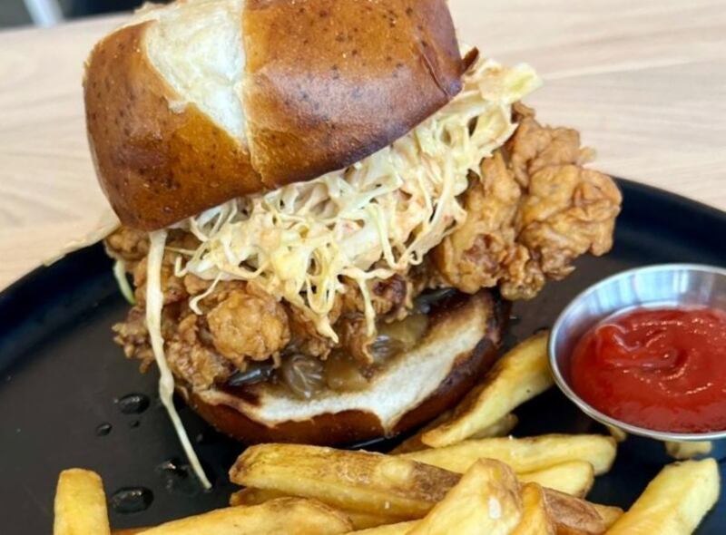Sándwich de pollo frito, servido en pan de pretzel con cebollas caramelizadas, ensalada de repollo con aderezo ranch y papas fritas.