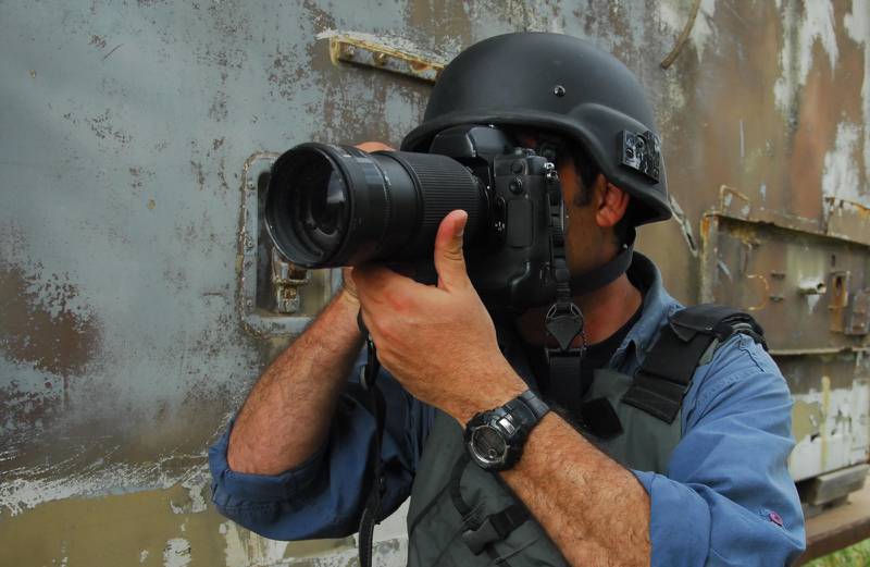 Un fotoperiodista apunta con el lente de su cámara en lo que parece ser una zona de conflicto. Lleva puesto un casco y chaleco.