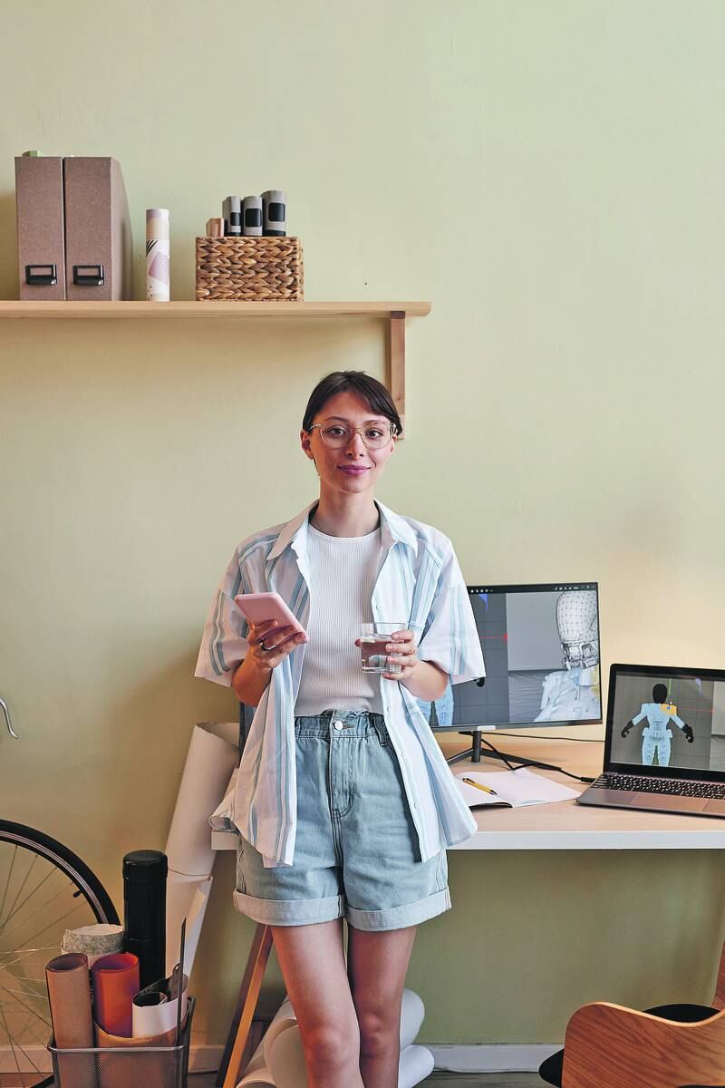 Fotografía de una mujer joven sonriendo a la cámara, con un celular en sus manos,  y a su lado una computadora que revela imágenes sobre impresiones 3d, lo que sugieres que es diseñadora en esta modalidad.
