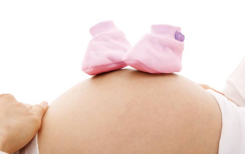 La panza de una mujer recostada, sobre la que descansan botines rosados de bebé.