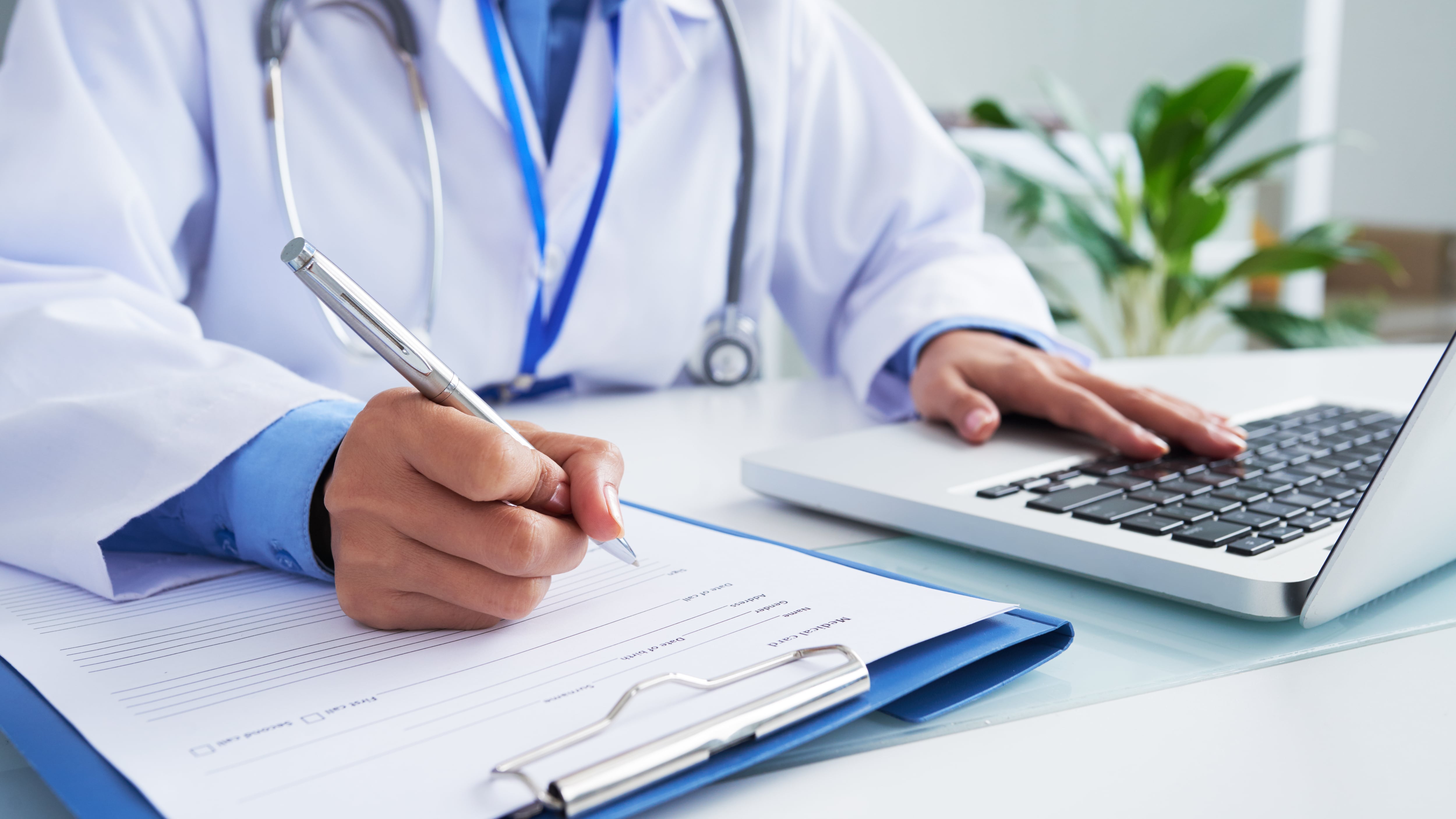 Manos de una doctora irreconocible escribiendo en formulario y escribiendo en el teclado de una computadora portátil