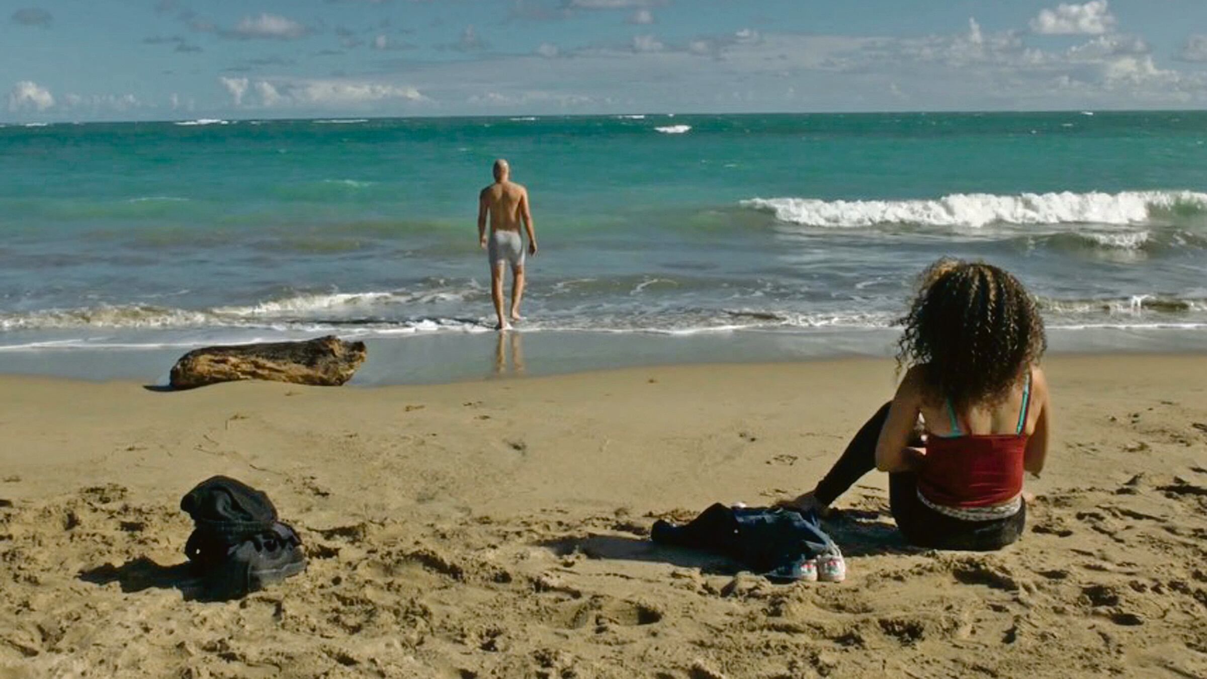 Una mujer aparece sentada en la arena, mirando hacia el mar, a la vez que un hombre camina a lo lejos en dirección del agua.
