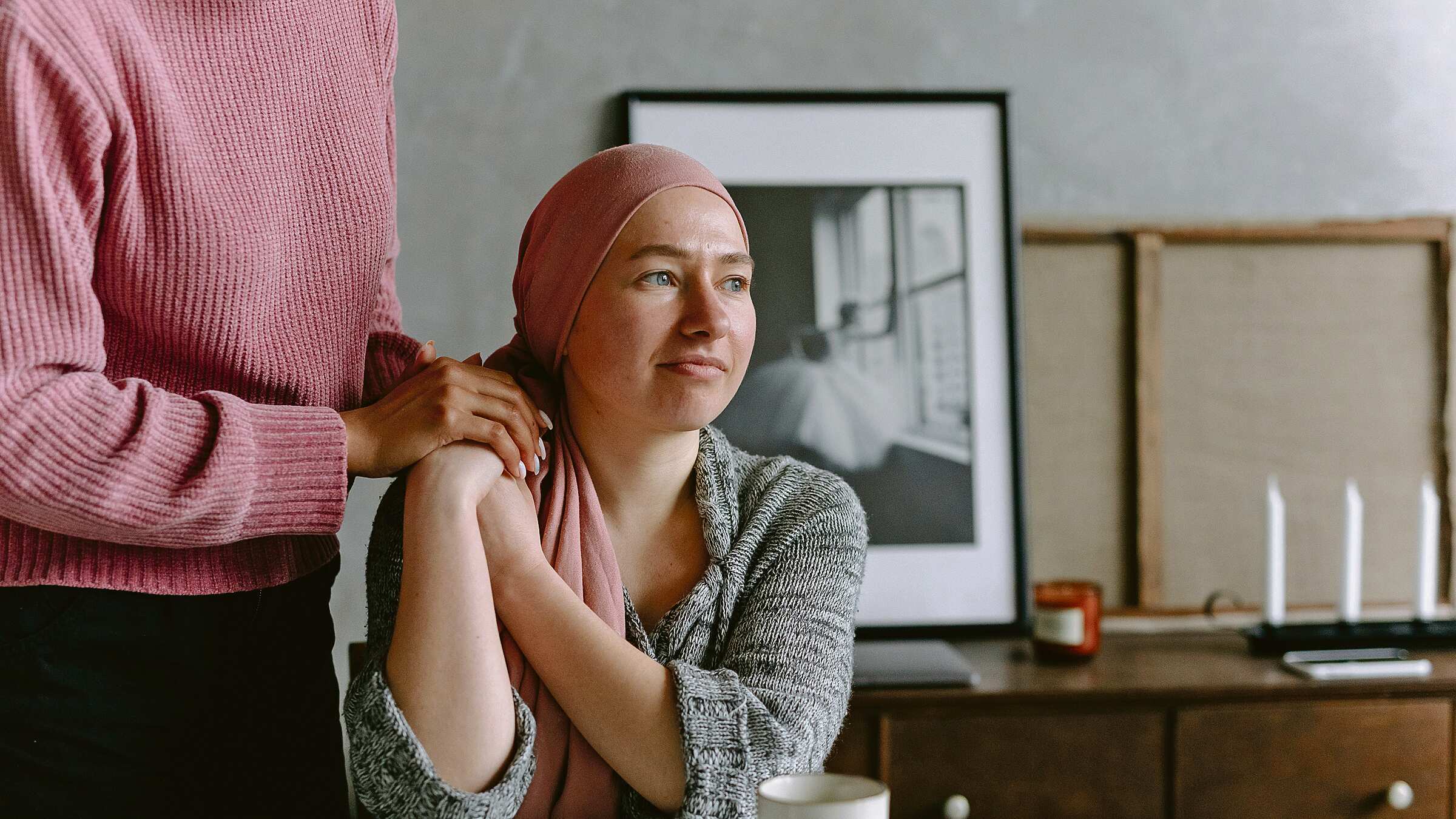 Una mujer con un turbante rosado, que aduce al cáncer de seno, está sentada frente a una mesa, mirando al horizonte, mientras una persona sostiene sus manos, que están recostadas en su hombro derecho.