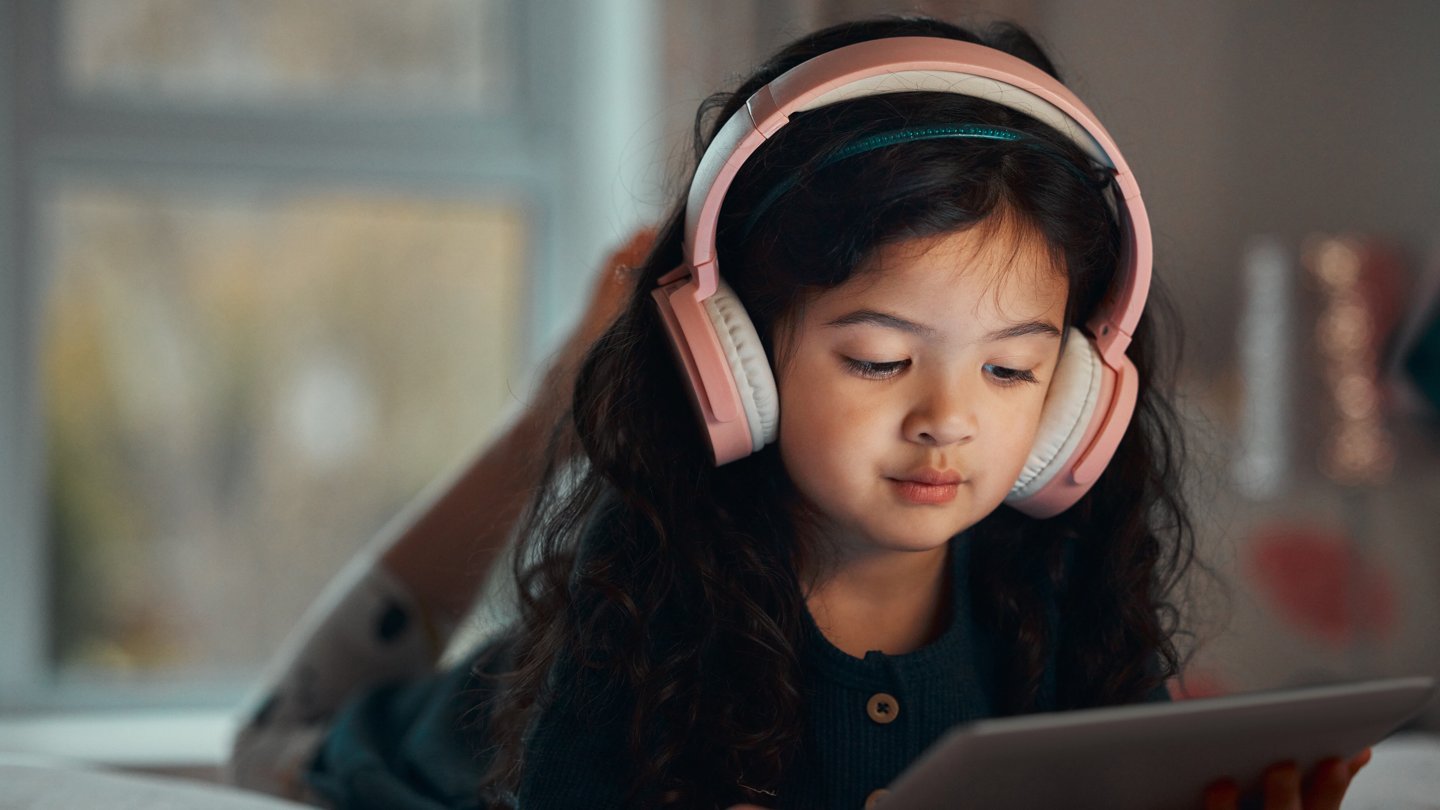 Una niña con audífonos auriculares rosados mira una tableta digital recostada en su cama.
