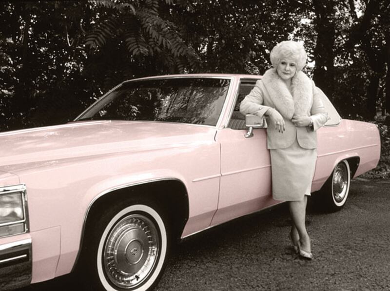 Mary Kay Ash posa frente a un Cadillac rosado en una imagen sepia con estética de los 60-70. Viste tacones, blazer y falda; un conjunto profesional.
