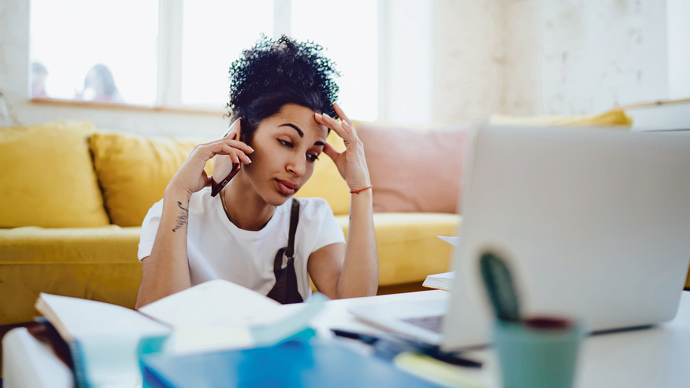 Una mujer sentada en el suelo de una sala, frente a una mesa repleta de papeles, una computadora portátil y otros materiales. Habla por teléfono y se lleva una mano a la frente. La imagen pretende mostrar a una trabajadora independiente.