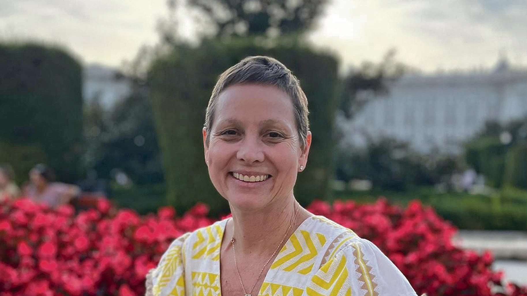 Saribel Matienzo, sobreviviente de cáncer de mama, mira a la cámara sonriendo frente a un arbusto con flores rosadas.