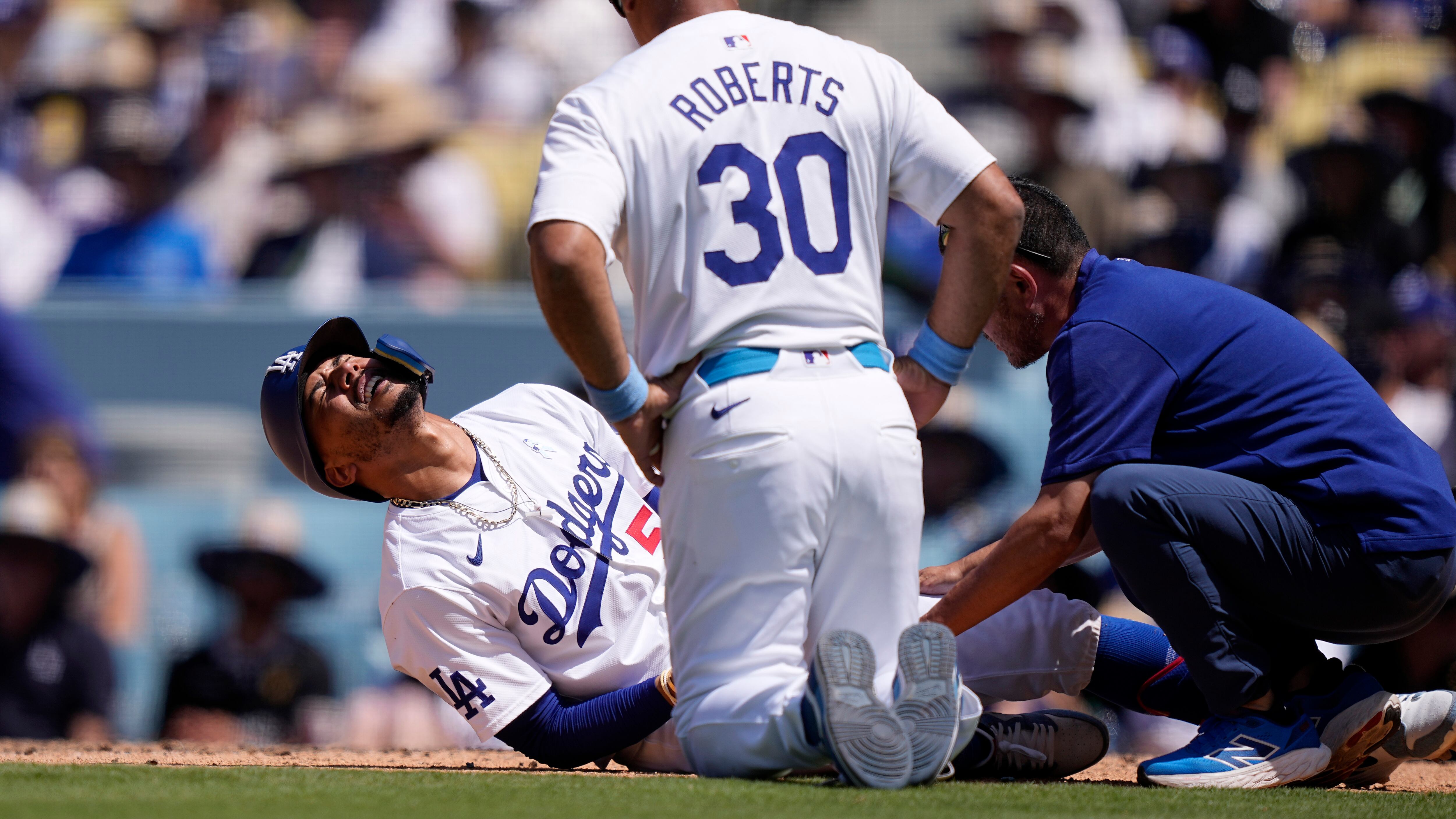 Mookie Betts, de los Dodgers de Los Ángeles (izquierda), se retuerce de dolor sobre el terreno tras ser golpeado por un lanzamiento, mientras que el mánager Dave Roberts y un entrenador lo asisten durante la séptima entrada del juego de béisbol ante los Reales de Kansas City, el 16 de junio de 2024, en Los Ángeles. (AP Foto/Mark J. Terrill)