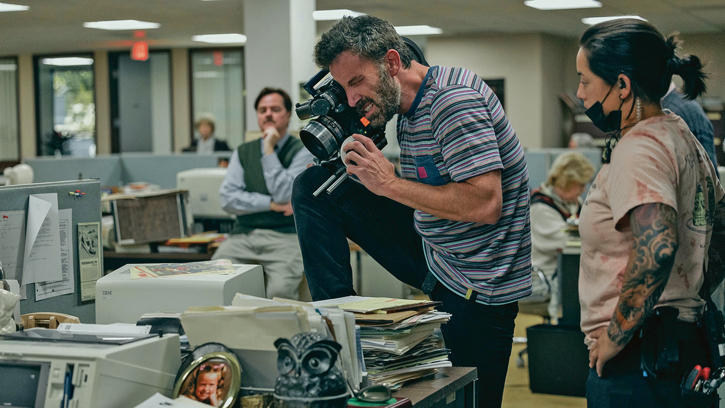 El director de Air, Ben Affleck, aparece con cámara en mano en el set, que incluye escritorios y un tono de oficina en los 80. Parte del equipo aparece en segundo plano.