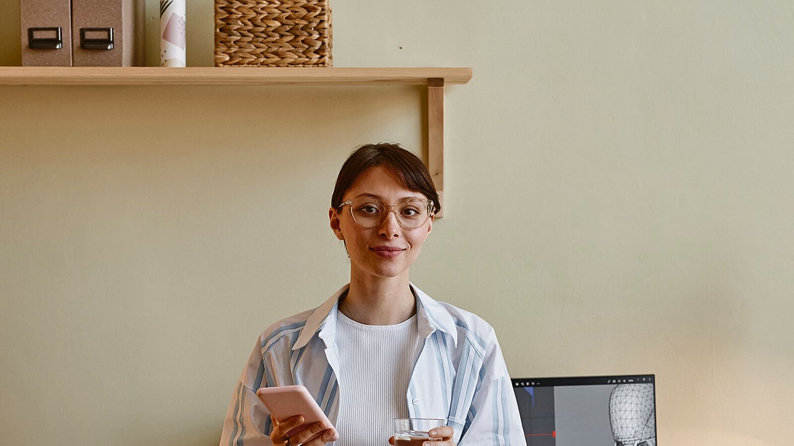 Fotografía de una mujer joven sonriendo a la cámara, con un celular en sus manos, y a su lado una computadora que revela imágenes sobre impresiones 3d, lo que sugieres que es diseñadora en esta modalidad.