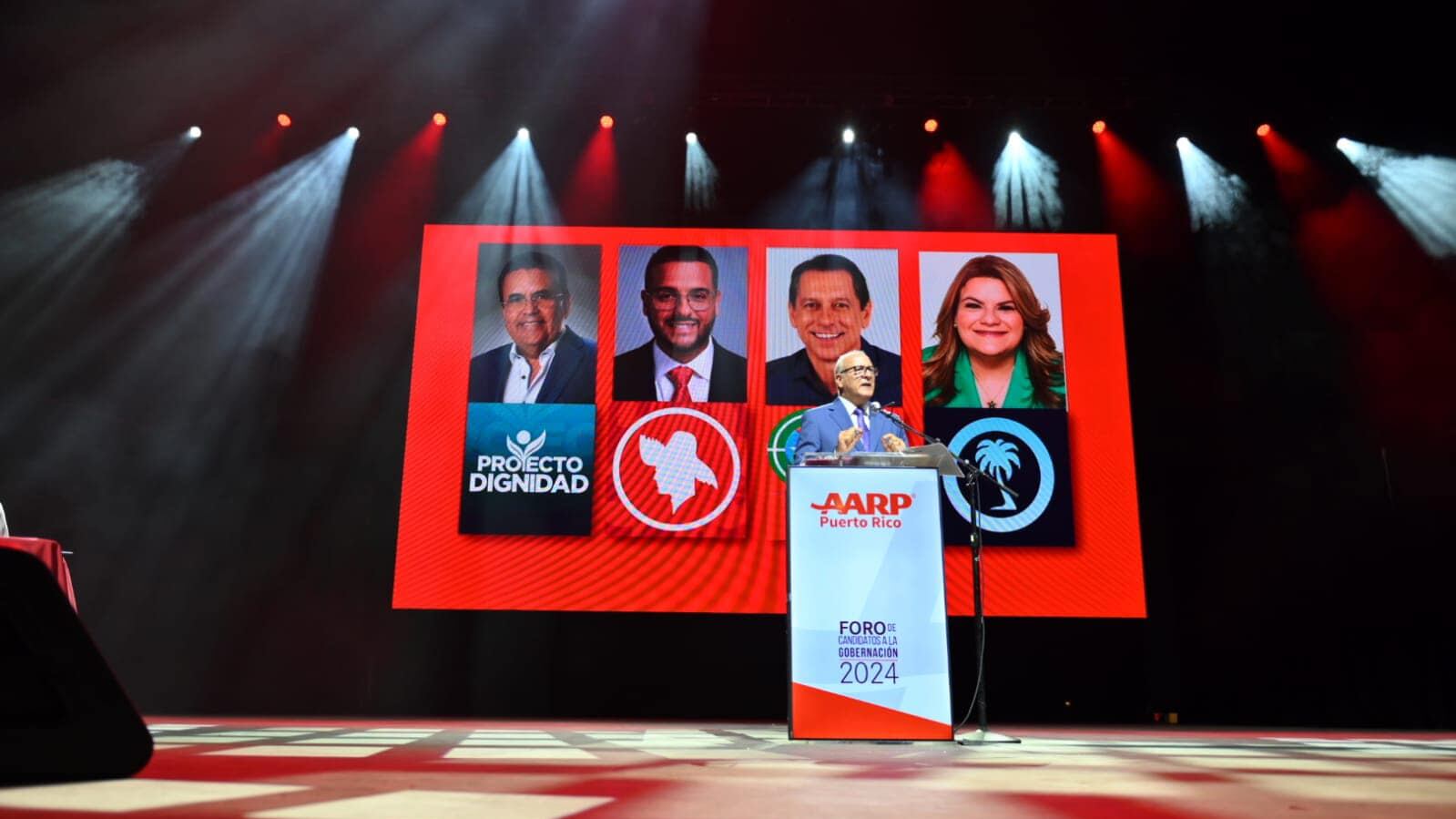Director estatal José Acarón frente a un podio con imagen de los cuatro aspirantes a la gobernación en la pantalla de fondo.