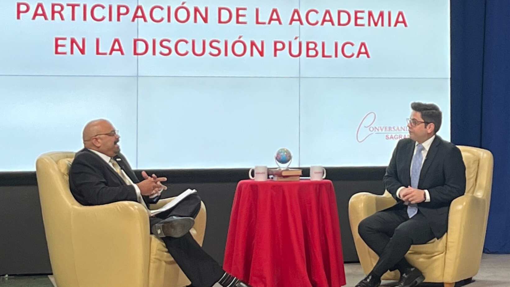 Rolando López y Francisco Parés sentados en un estudio. Al fondo, una pantalla lee "Participación de la academia en la discusión pública".