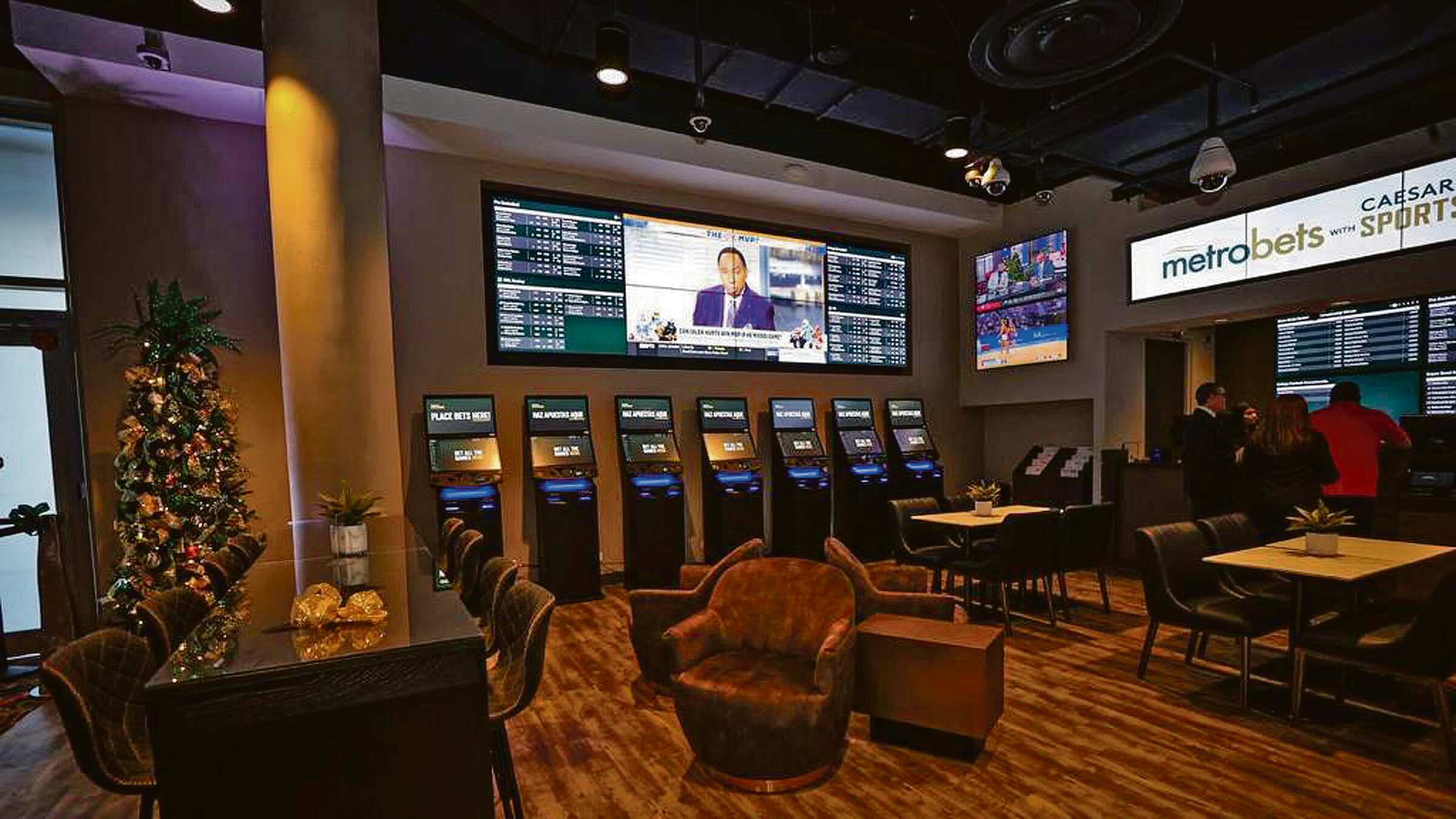 En una sala de apuestas, MetroBets, una línea de máquinas de juego y varias pantallas rellenan la pared de fondo, mientras que un grupo de personas se acercan al mostrador.