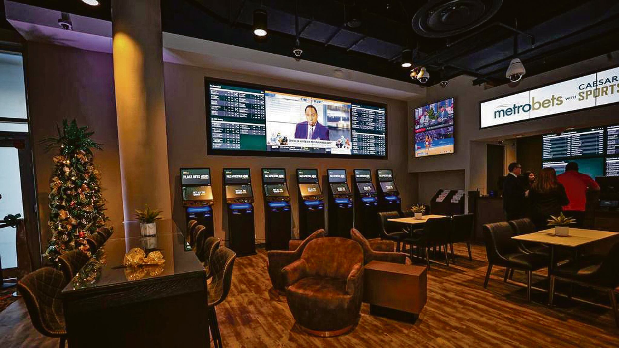 En una sala de apuestas, MetroBets, una línea de máquinas de juego y varias pantallas rellenan la pared de fondo, mientras que un grupo de personas se acercan al mostrador.