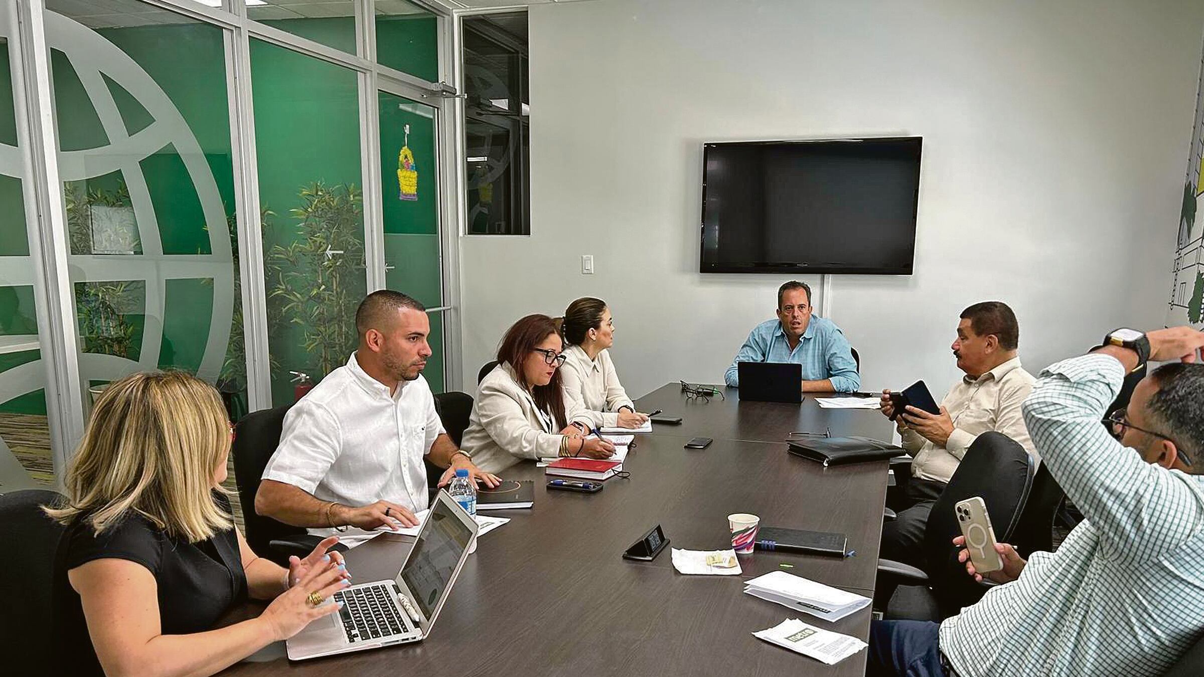Los periodistas Aiola Virella y Rafael Lenín López en una mesa junto a cinco personas, de los candidatos del PPD, en una oficina con los colores verde y blanco, propios de la marca de Metro.